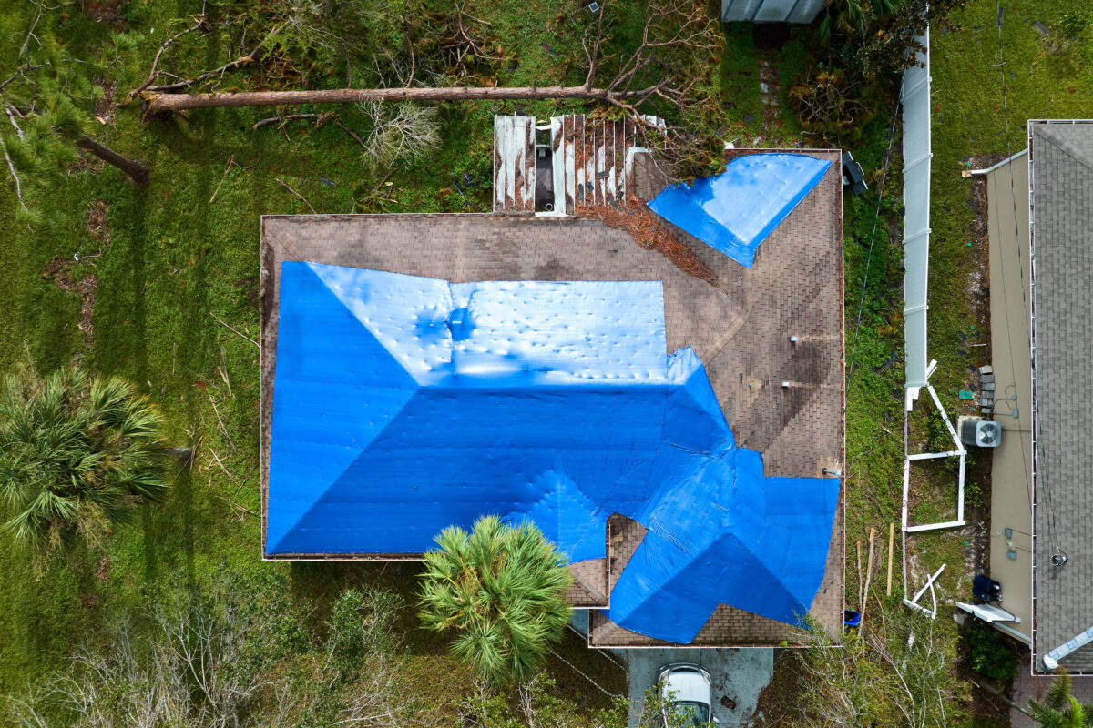 Aerial view of damaged in hurricane Ian house roof covered with blue protective tarp against rain water leaking until replacement of asphalt shingles.